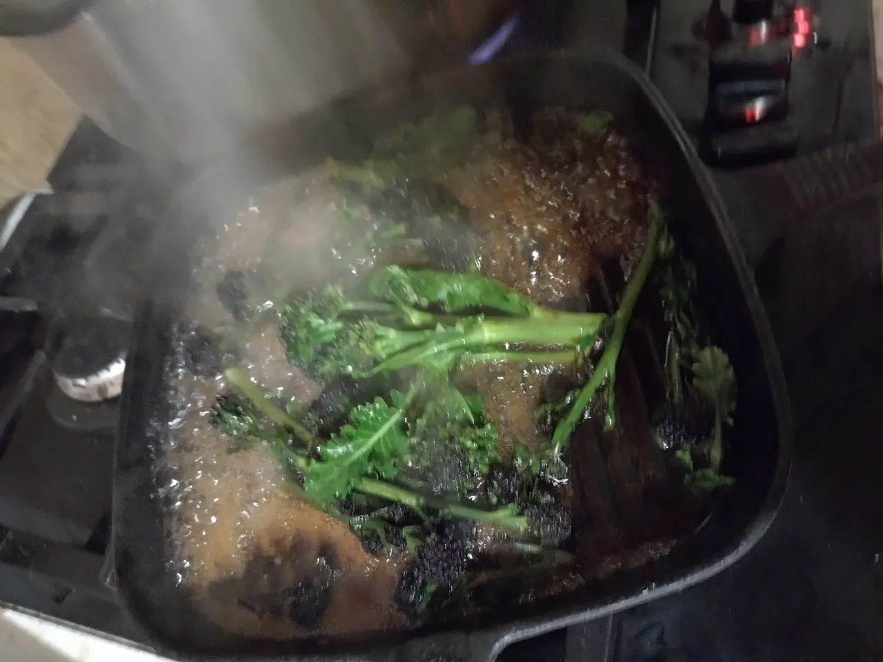 Purple sprouting broccoli braising in butter, soy sauce, and lemon juice with steam rising from the griddle pan