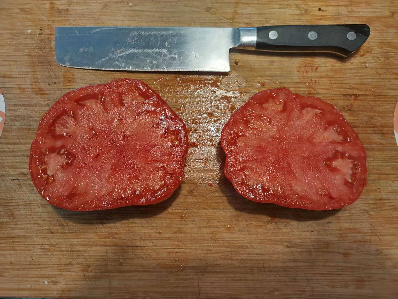 A large beefsteak tomato sliced in half on a wooden chopping board with a knife, showing the dense fleshy interior with minimal seeds