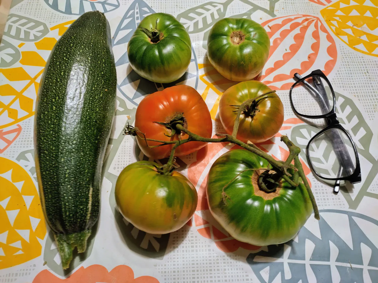Five large heirloom tomatoes in green, orange, and red alongside a courgette and glasses for scale on a colourful tablecloth