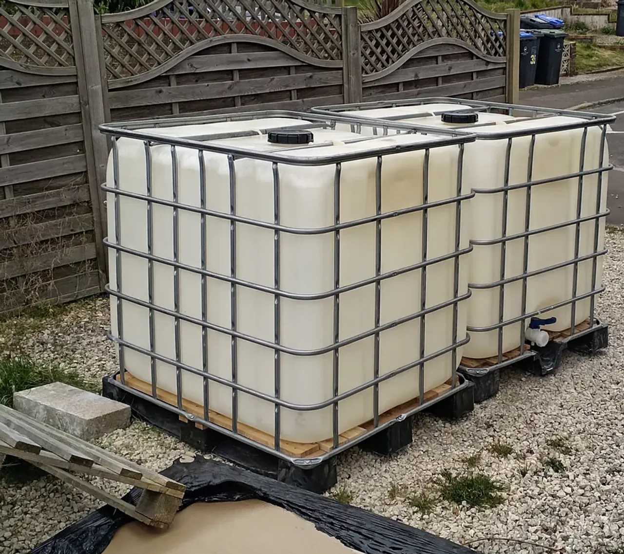 Two white IBC tote containers in metal cage frames on gravel, freshly delivered for the hydroponic system