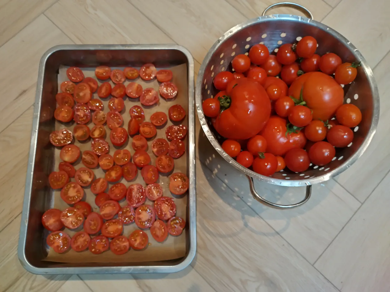 Tray of halved cherry tomatoes ready for roasting alongside a colander full of fresh whole tomatoes in mixed sizes