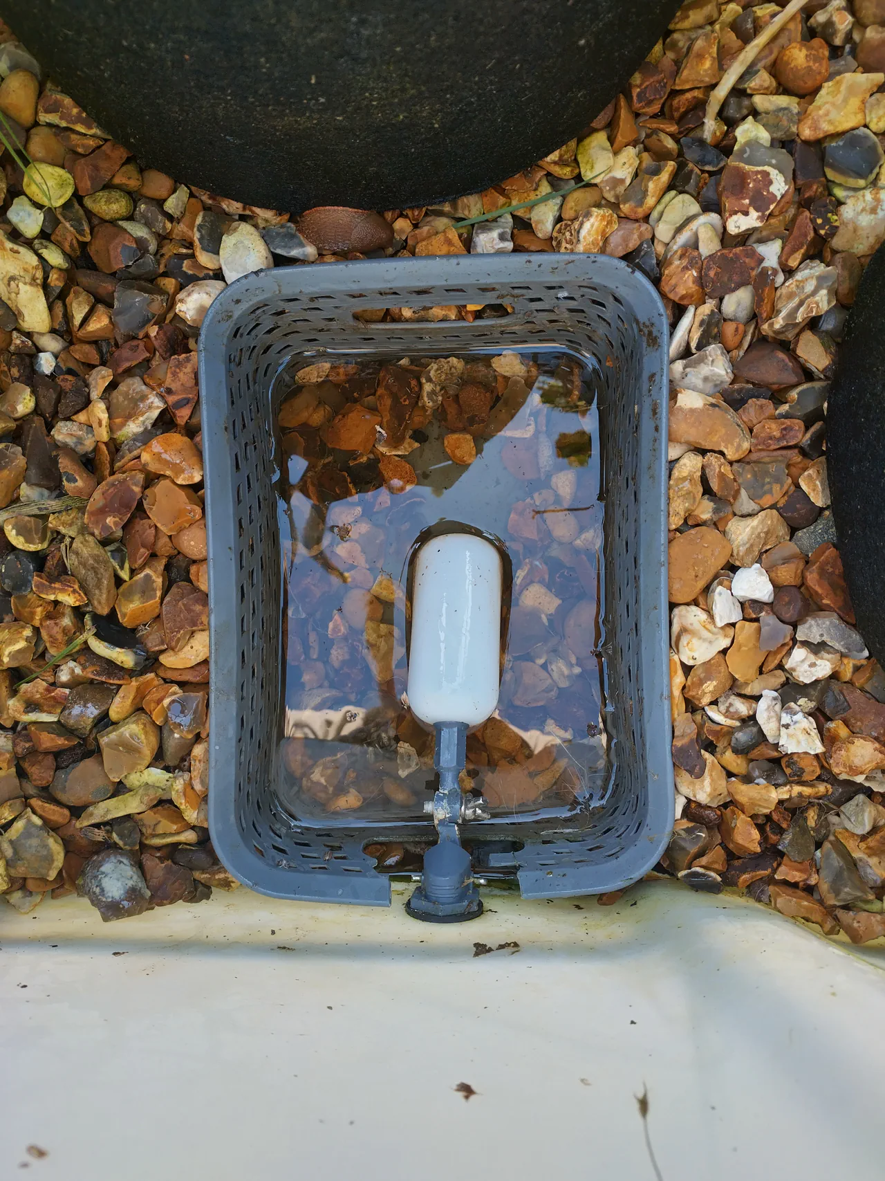 The float valve chamber: a plastic cutlery basket from Tesco, with the float valve visible inside and the water level just below the gravel surface