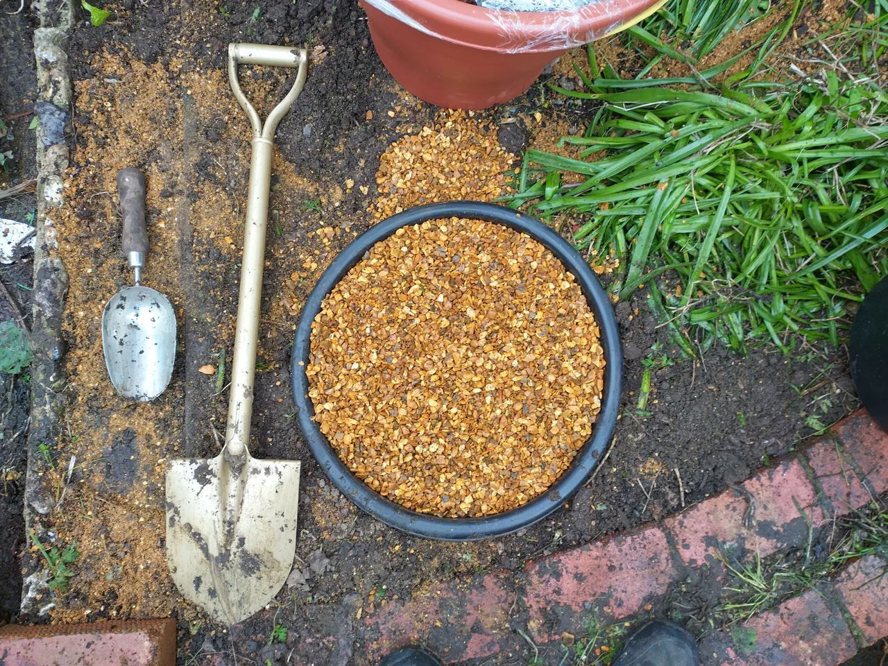 The black centre pot sitting on a gravel base with a spade alongside, showing the level gravel pad prepared for the planter